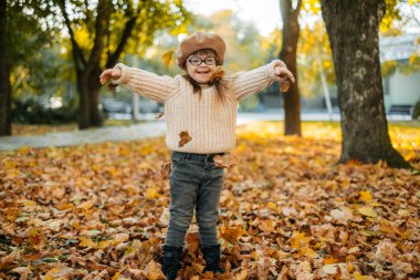 Mutlu, kahverengi saçlı, örgü örmüş bir kadın, gökyüzüne parlak sonbahar yaprakları fırlatıyor ve gülüyor, çocuk sıcak bir sonbahar parkında eğleniyor, mutlu bir çocukluk geçiriyor.