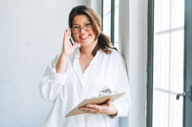 Genç gülümseyen esmer kadın, pencerenin yanında beyaz gömlekli, parlak modern ofiste elinde kalem ve belgelerle.
