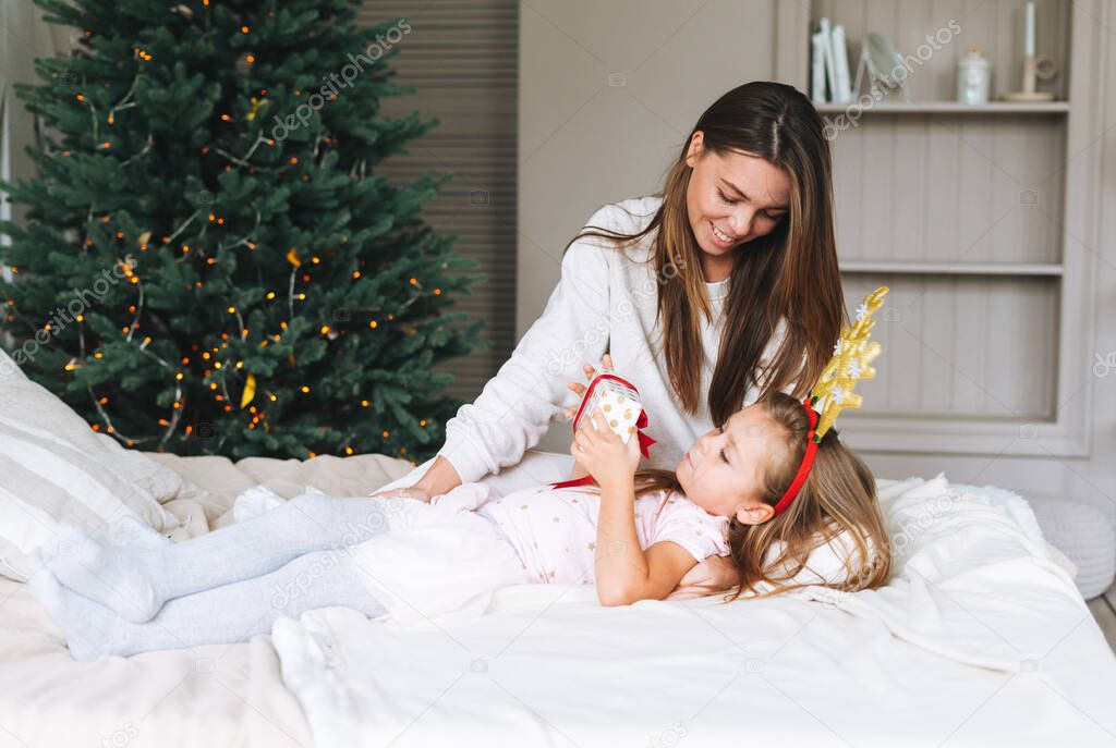 Cute funny little girl with mother on bed in room with Christmas tree. Portrait of child in the rim with deer horns in cottage house in christmas time, happy New Year. Happy family presents christmas gifts