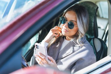 Uzun sarı saçlı, güneş gözlüklü, araba süren ve cep telefonu kullanan şık bir kadın.
