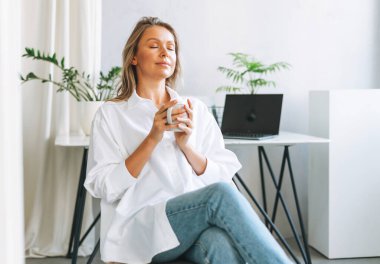 Uzun saçlı, beyaz tişörtlü, gülümseyen genç sarışın kadın dinleniyor ve modern ofiste çay içiyor.