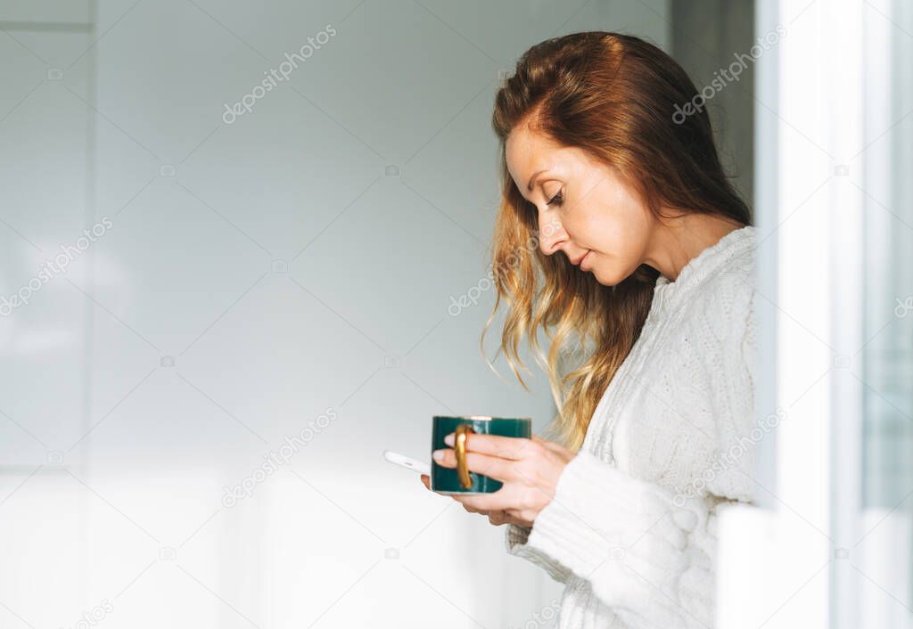 Young adult forty years beautiful blonde woman with long hair using smartphone and drinking morning tea on kitchen at home