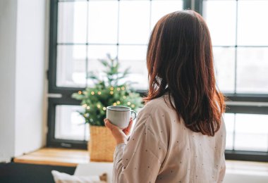 40 yaşında, genç, siyah saçlı, rahat pijamalı bir kadın oturma odasında bir fincan çayla Noel ağacının arkasından insanlar.