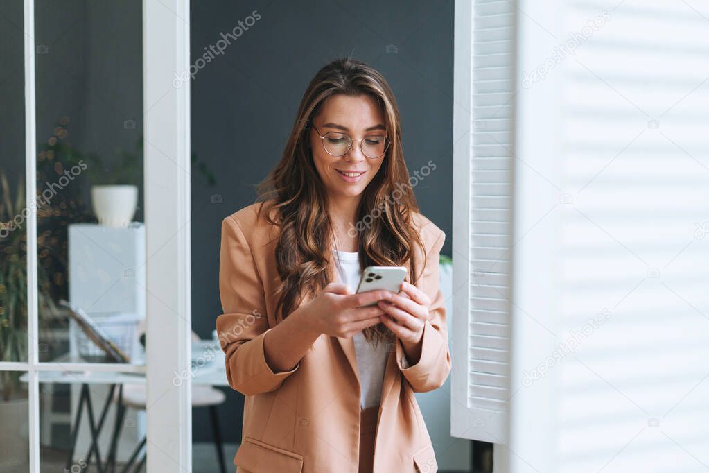 Young smiling brunette business woman in glasses with long hair in stylish beige suit using smartphone in modern office