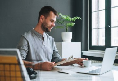 Gündelik gri uzun kollu, gülümseyen genç iş adamı karanlık modern ofis odasında dizüstü bilgisayarda çalışıyor.