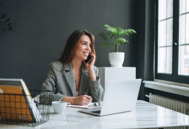 Gri takımlı uzun saçlı, gülümseyen esmer bir kadın cep telefonu kullanıyor ve modern ofiste dizüstü bilgisayarla çalışıyor.