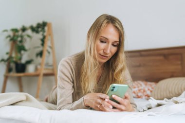 Evdeki yatakta cep telefonu kullanan güzel sarışın genç kadın.