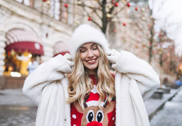Beyaz kürklü, kıvırcık saçlı, kışın Noel panayırında ışıklar altında eğlenen genç ve mutlu bir kadın.