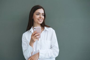 Portrait of beautiful smiling woman student with dark long hair in white shirt using mobile phone in the hand on grey background isolated, generation Z
