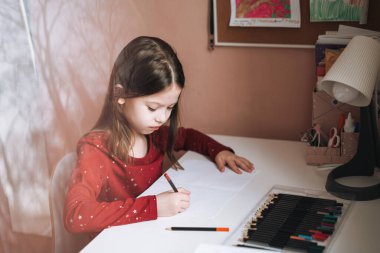 Kırmızı elbiseli, uzun saçlı güzel bir kız çocuk odasında resim çiziyor.