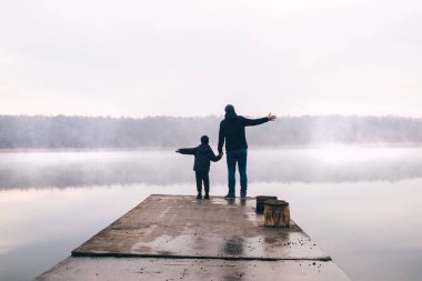 Baba ve oğlu sisli bir dağ gölünde. Sihirli sabah. Sis su yüzeyine güzel bir şekilde yayılır. Ağaçlar neredeyse görünmez. Bulutlar ve gökyüzü yüzeyden yansıyor. Balıkçı iskelesi arkaplanı