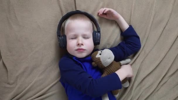 ヘッドフォンの男の子は寝る前に子供のための瞑想を聞く 子供の健康的な神経系の概念 寝る前のリラクゼーション 金髪の男が家でリラックスしている 聴覚療法 — ストック動画