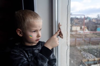 Rus saldırısı Ukrayna 'da. Çocuk pencereye oturur ve ağlar. Yıkık bir binanın arka planında çatlaklar ve yıkım var. Çatışmaların ve sivillerin bombalanmasının sonuçları