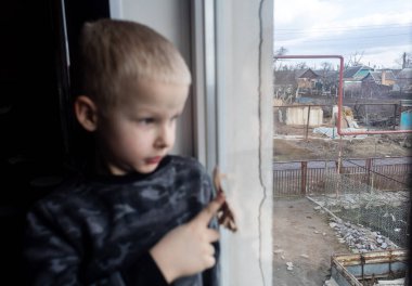 Rus saldırısı Ukrayna 'da. Çocuk pencereye oturur ve ağlar. Yıkık bir binanın arka planında çatlaklar ve yıkım var. Çatışmaların ve sivillerin bombalanmasının sonuçları
