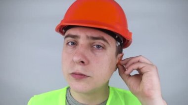 Başlık takan ve yeşil yelek giyen bir işçi kulak tıkacı takar. Endüstriyel güvenlik. Endüstriyel gürültü koruması. Gürültülü çalışma kurallarına uymak. Turuncu kulak tıkaçları