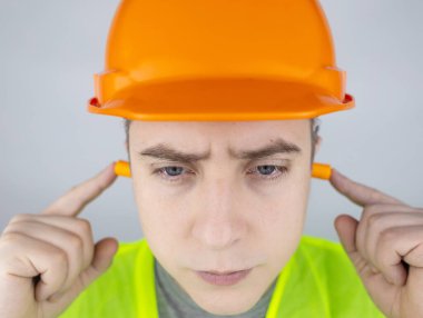 Başlık takan ve yeşil yelek giyen bir işçi kulak tıkacı takar. Endüstriyel güvenlik. Endüstriyel gürültü koruması. Gürültülü çalışma kurallarına uymak. Turuncu kulak tıkaçları