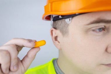 Başlık takan ve yeşil yelek giyen bir işçi kulak tıkacı takar. Endüstriyel güvenlik. Endüstriyel gürültü koruması. Gürültülü çalışma kurallarına uymak. Turuncu kulak tıkaçları