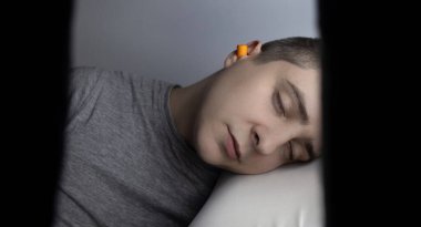 Adam kulak tıkacı takıyor. Turuncu gürültü bariyerine yakın çekim. Derin uykuda. KBB doktoru çevredeki gürültüyü azaltmak için turuncu kulaklık tavsiye ediyor. Seslere karşı hassasiyet artışı.