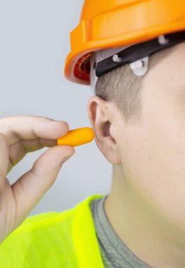 Başlık takan ve yeşil yelek giyen bir işçi kulak tıkacı takar. Endüstriyel güvenlik. Endüstriyel gürültü koruması. Gürültülü çalışma kurallarına uymak. Turuncu kulak tıkaçları