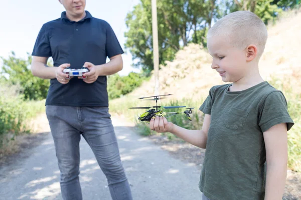 Dad and child playing with radio-controlled helicopter. Blond boy holds in his hand a mini-model of a helicopter in which propellers work. Kid develops fine motor skills. Happy childhood and love