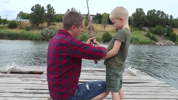 父と息子の釣り 父は息子に回転を保持し リールを回転させる方法を示します 池や川での釣りの訓練 親の考え方 — ストック動画