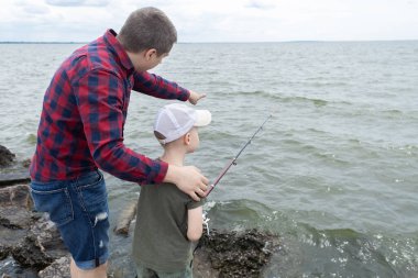 Baba oğul balık tutuyor. Baba oğluna makarayı nasıl tutup döndüreceğini gösteriyor. Gölde ya da nehirde balık tutma eğitimi. İlgili ebeveyn kavramı.