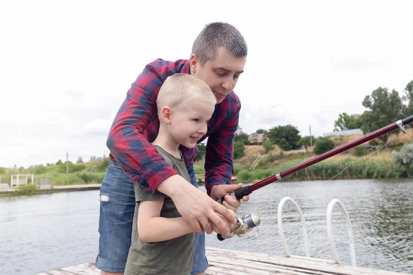 Father and son fishing. Dad shows his son how to hold the spinning and spin the reel. Fishing training on a pond or river. Caring parent concept.