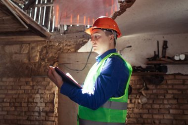 Building inspector. Man in a hard hat and a yellow reflective vest examines damaged structures and inspects the building. Damage assessment. Preparing for the repair or construction of a building