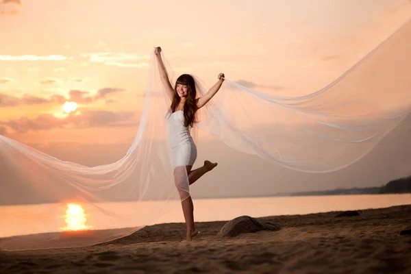 Bela noiva com um longo véu na praia ao pôr do sol — Fotografia de Stock