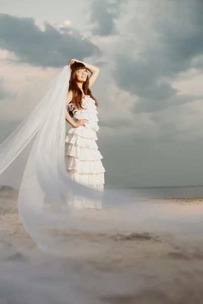 Schöne Braut mit langem Schleier am Strand bei Sonnenuntergang — Stockfoto