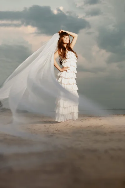 Όμορφη νύφη με ένα μακρύ πέπλο πάνω στην παραλία στο ηλιοβασίλεμα Εικόνα Αρχείου