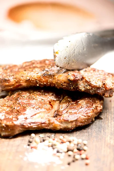 Grilled steaks on an old wooden board with tongs — Stock Photo, Image