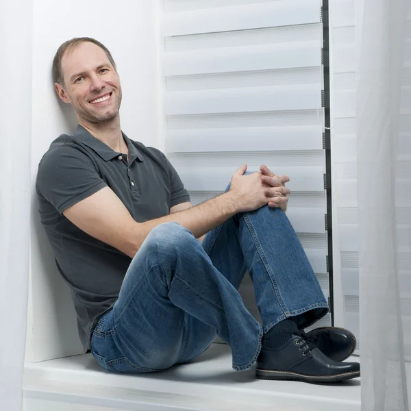 Młody człowiek uśmiechający się siedząc na białe okna — Zdjęcie stockowe