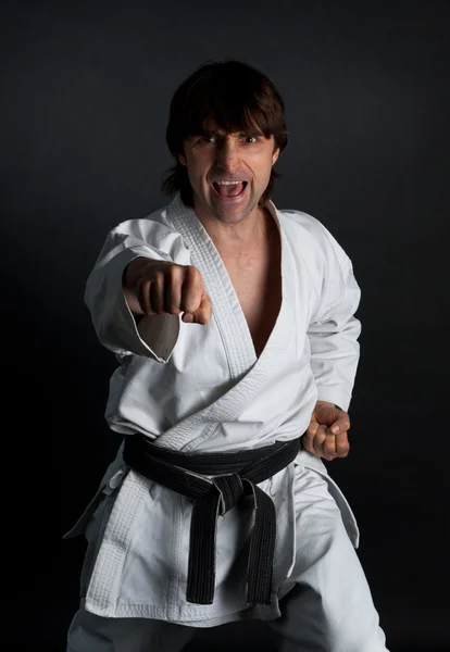 Man in a kimono with a black belt doing  forward kick on a black — Stock Photo, Image