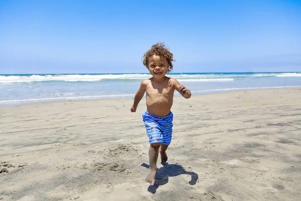 家族の休暇中にアフリカ系アメリカ人の少年が走ってビーチで遊んでいるのを笑っています 海と砂の中で遊んで楽しく活動している — ストック写真