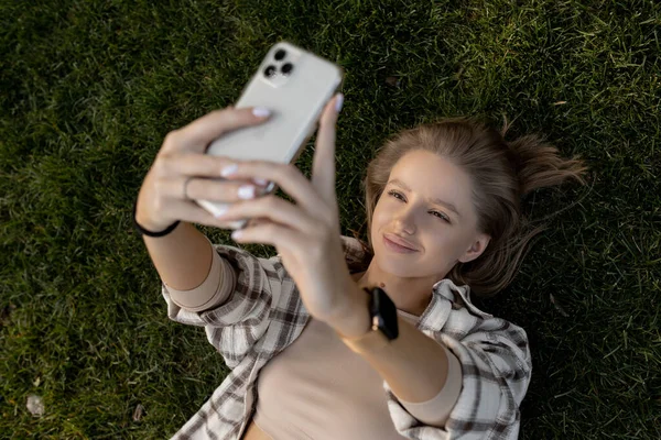 여자가 초록빛 풀밭에 누워 전화기를 들여다보며 셀카를 만듭니다. — 스톡 사진