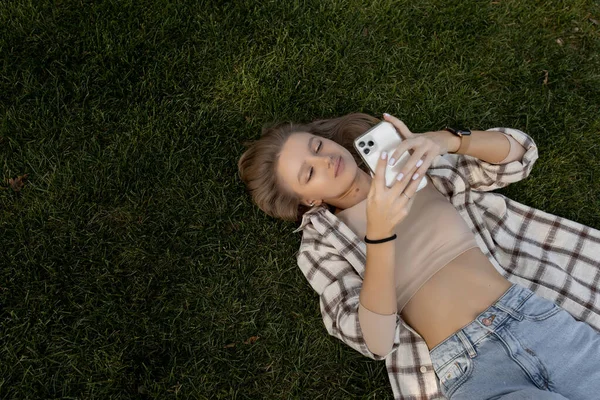 Молодая женщина лежит на зеленой траве и улыбается, глядя в телефон — стоковое фото
