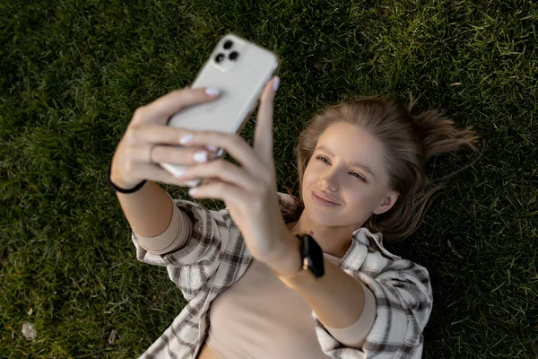 Mujer se encuentra en la hierba verde mientras mira en el teléfono, hace una selfie Fotos De Stock