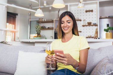 Evde dinlenen, cep telefonu kullanan ve bir bardak şarap içen genç bir kadın..