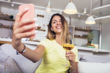 Genç bir kadın evde dinleniyor, cep telefonuyla selfie çekiyor ve bir bardak şarap içiyor..