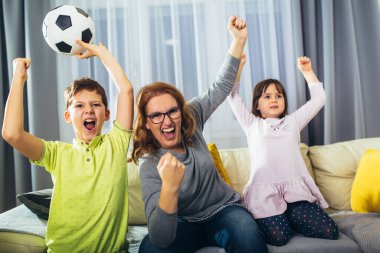 Anne ve çocukları kollarını kaldırıp çığlık atıyorlar. Futbol maçını seyredip evdeki golü kutluyorlar.