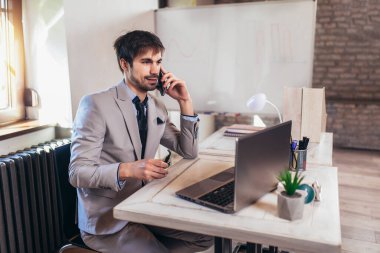 Genç işadamı ofiste dizüstü bilgisayar ve telefon kullanıyor.