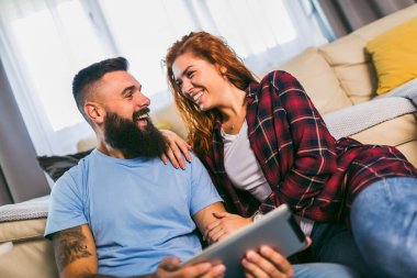 Genç bir çift, oturma odasındaki tablette medyanın içeriğini internetten izliyor.