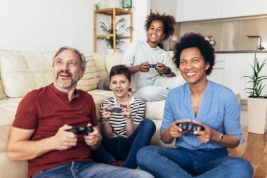 Gülümseyen aile evde birlikte vakit geçiriyorlar. Oturma odasında kanepede oturup video oyunları oynuyorlar..