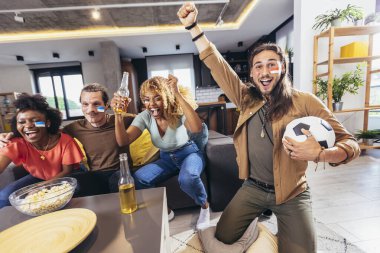 Young group watching sports on television and cheering. Emotional football fans watching football game together at home. Sports Fans with Painted Faces Sitting on a Couch Watch Game on TV