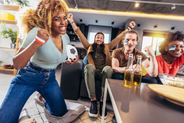 Young group watching sports on television and cheering. Emotional football fans watching football game together at home. Sports Fans with Painted Faces Sitting on a Couch Watch Game on TV