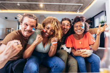 Young group watching sports on television and cheering. Emotional football fans watching football game together at home. Sports Fans with Painted Faces Sitting on a Couch Watch Game on TV