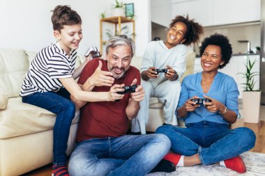 Gülümseyen aile evde birlikte vakit geçiriyorlar. Oturma odasında kanepede oturup video oyunları oynuyorlar..