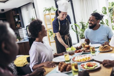Yemek masasında yemek yiyen çok nesilli Afro-Amerikan bir aile. Beyaz kadın aşçı servis ediyor..