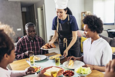 Yemek masasında yemek yiyen çok nesilli Afro-Amerikan bir aile. Beyaz kadın aşçı servis ediyor..